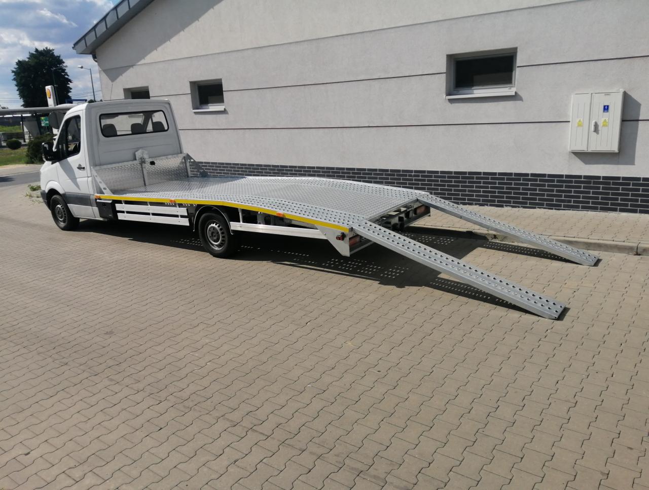 Biała laweta z rozłożonymi najazdami na brukowanym placu, z budynkiem w tle.