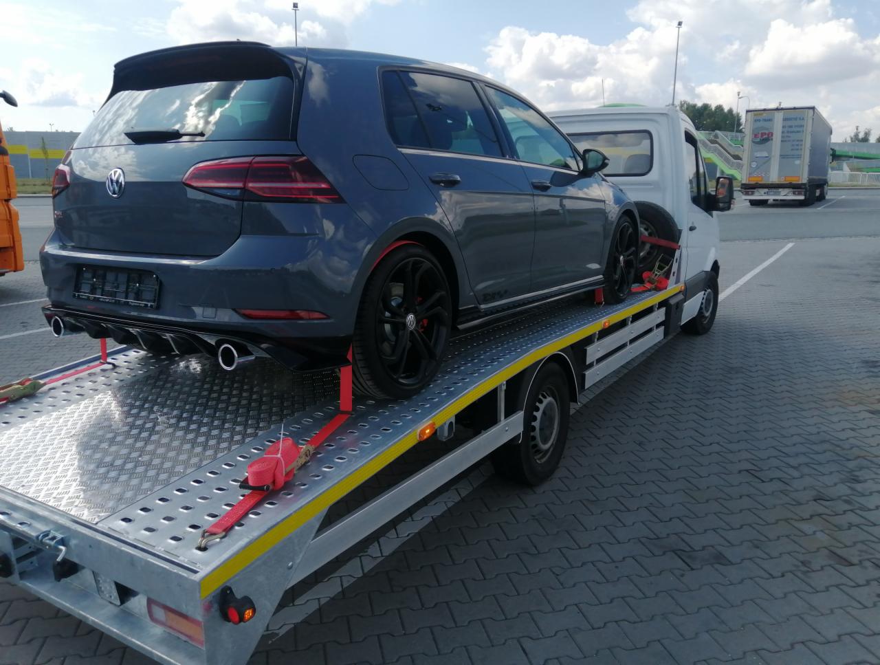 Szary samochód Volkswagen Golf na platformie białej lawety, zabezpieczony czerwonymi pasami transportowymi, na parkingu z kostki brukowej, w tle biała ciężarówka.