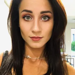 Portret kobiety z ciemnymi włosami, delikatnym makijażem i srebrnym naszyjnikiem z krzyżykiem, patrzącej wprost w obiektyw. W tle widoczny fragment wnętrza.