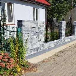 Nowoczesne ogrodzenie z szarej kostki imitującej kamień i czarnych, kutych przęseł, widoczne z perspektywy chodnika, z elementami zieleni w tle i kwiatami w pierwszym planie.