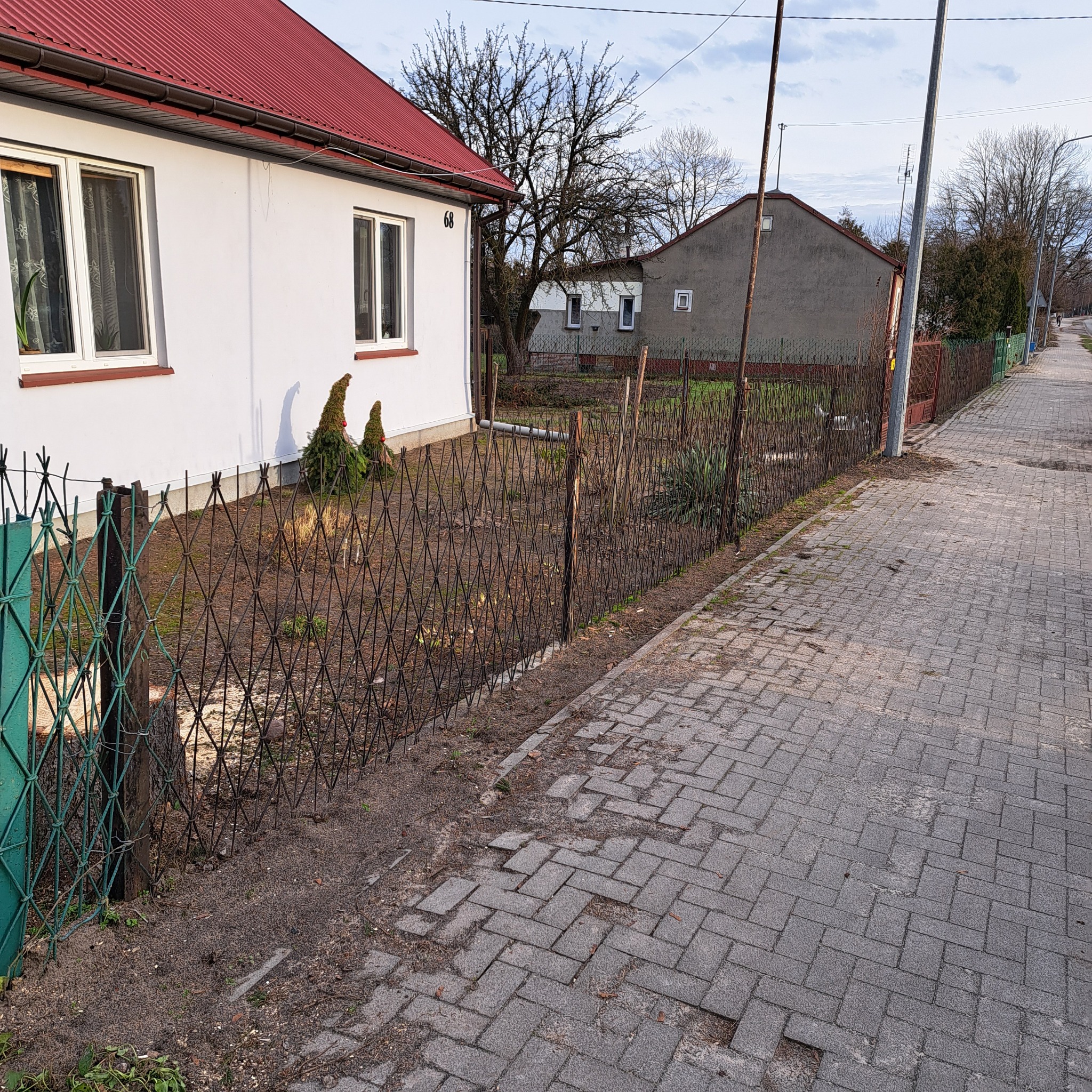 Niski płot z siatki rombowej, oddzielający posesję z białym domem i czerwonym dachem od chodnika z kostki brukowej.
