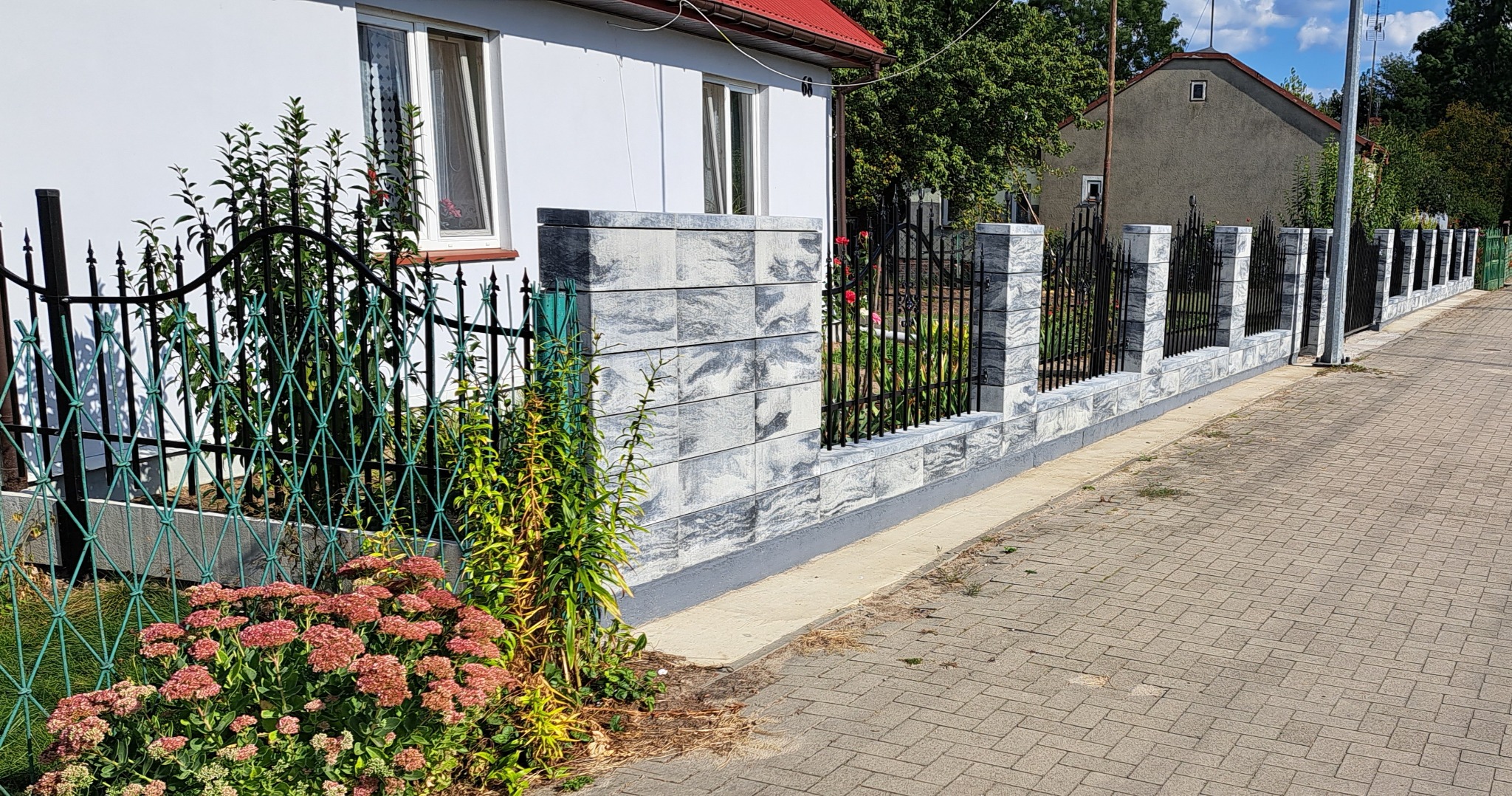 Nowoczesne ogrodzenie z szarej kostki imitującej kamień i czarnych, kutych przęseł, widoczne z perspektywy chodnika, z elementami zieleni w tle i kwiatami w pierwszym planie.