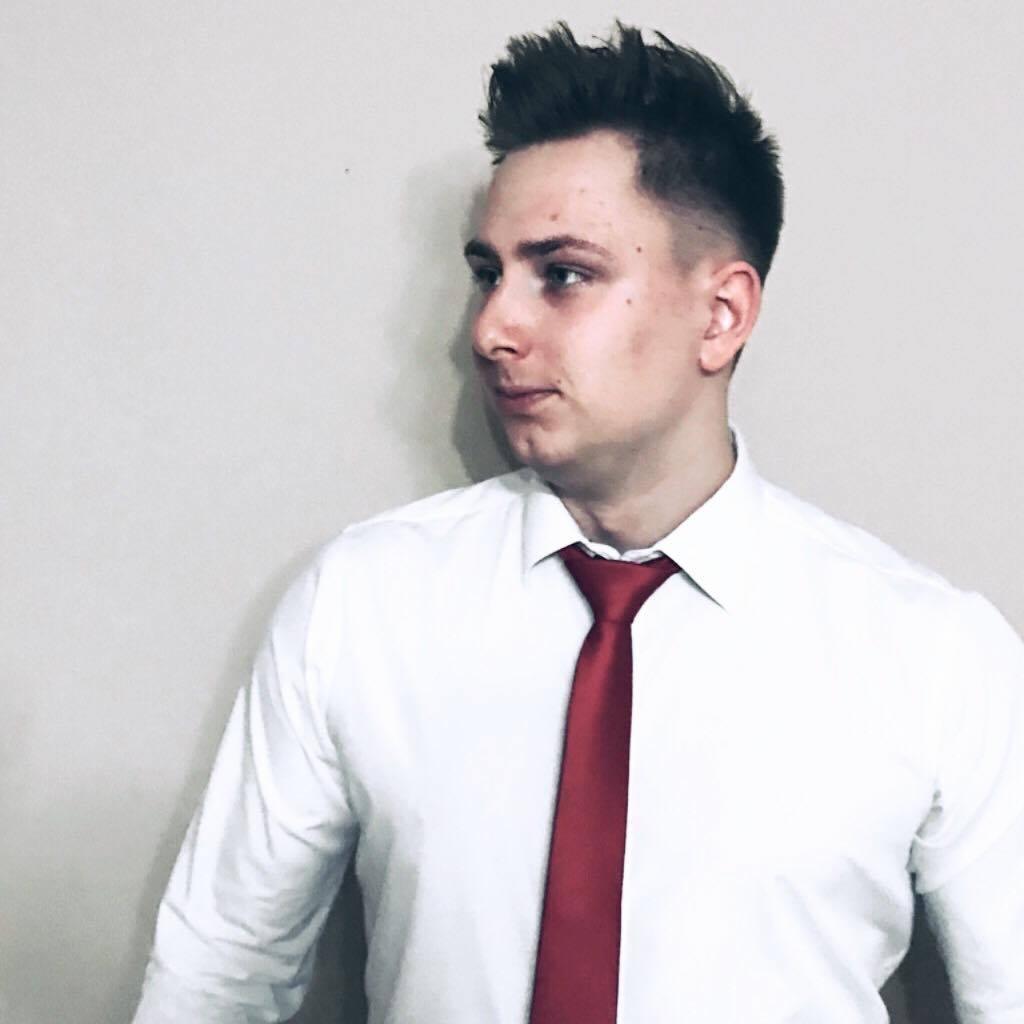 Młody mężczyzna w białej koszuli i czerwonym krawacie, patrzy w lewo, na jasnym tle. Portret biznesowy z charakterystyczną fryzurą.