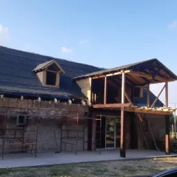 Dom w trakcie przebudowy z widocznym rusztowaniem, nową konstrukcją balkonu i częściowo pokrytym dachem, uchwycony w słoneczny dzień.