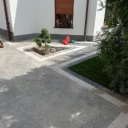 Ułożona szara kostka brukowa z białymi obramowaniami wokół trawnika i drzewka, zagęszczarka w tle, kwiaty w oknie budynku.