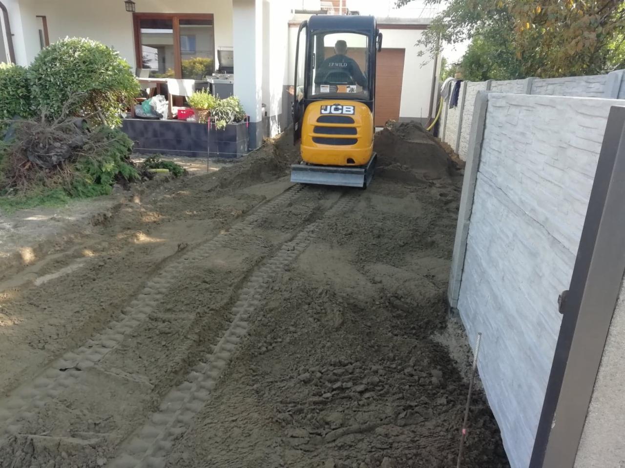 Żółta mini koparka JCB wyrównuje teren przy betonowym ogrodzeniu, widoczne ślady opon w piasku.