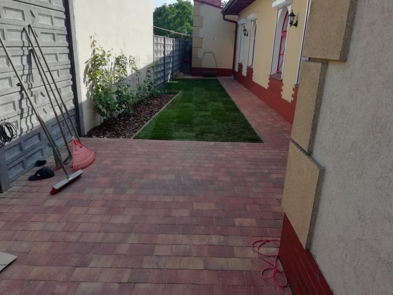 Ułożona czerwona kostka brukowa na posesji, widoczny trawnik, budynek z oknami i narzędzia ogrodnicze przy ścianie.