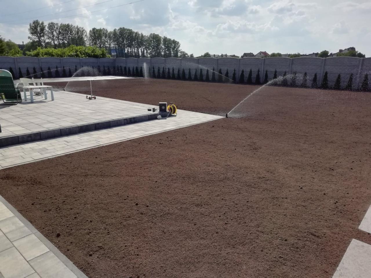 Nawadnianie wyrównanego terenu pod trawnik z systemem zraszaczy obok ułożonej kostki brukowej.