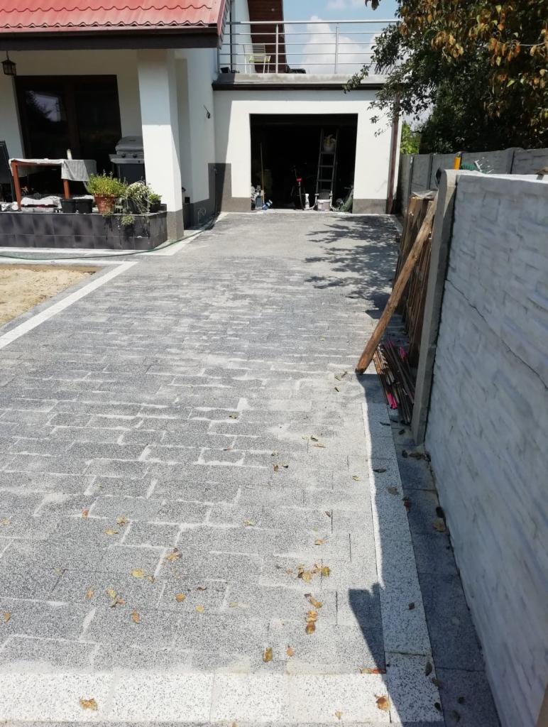 Świeżo ułożona, szara kostka brukowa prowadząca do garażu domu jednorodzinnego, z widocznymi detalami wykończenia i kilkoma opadłymi liśćmi.