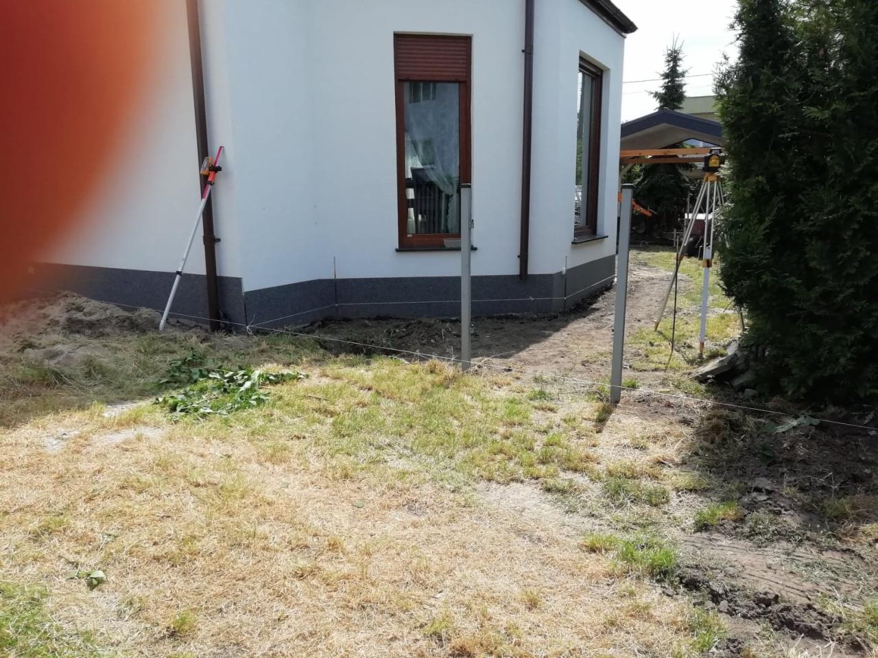 Przygotowanie terenu pod budowę domu: wytyczone granice, wbite słupki, niwelator laserowy na statywie, trawa częściowo uschnięta.