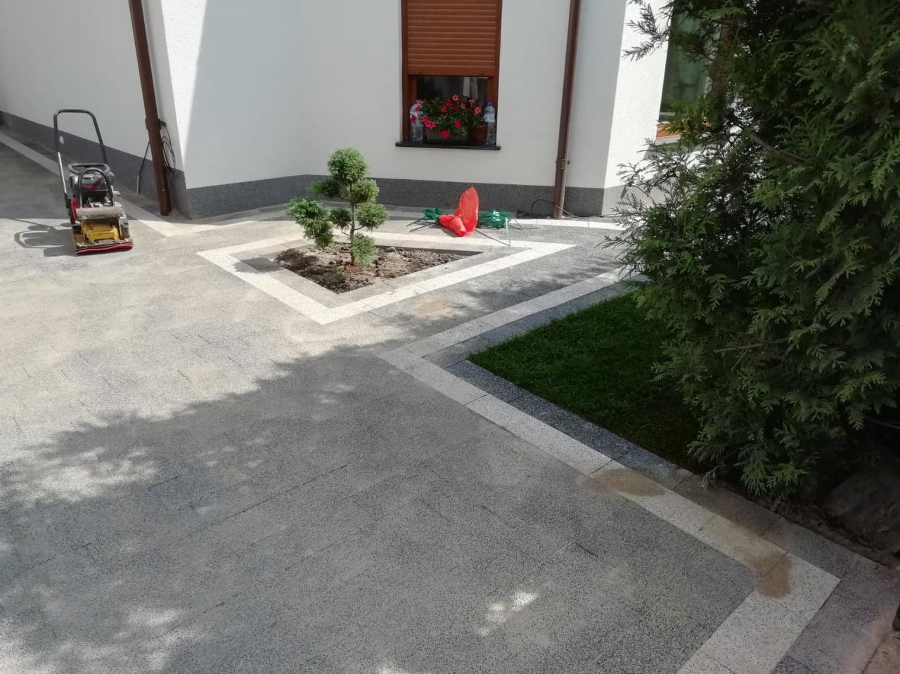 Ułożona szara kostka brukowa z białymi obramowaniami wokół trawnika i drzewka, zagęszczarka w tle, kwiaty w oknie budynku.