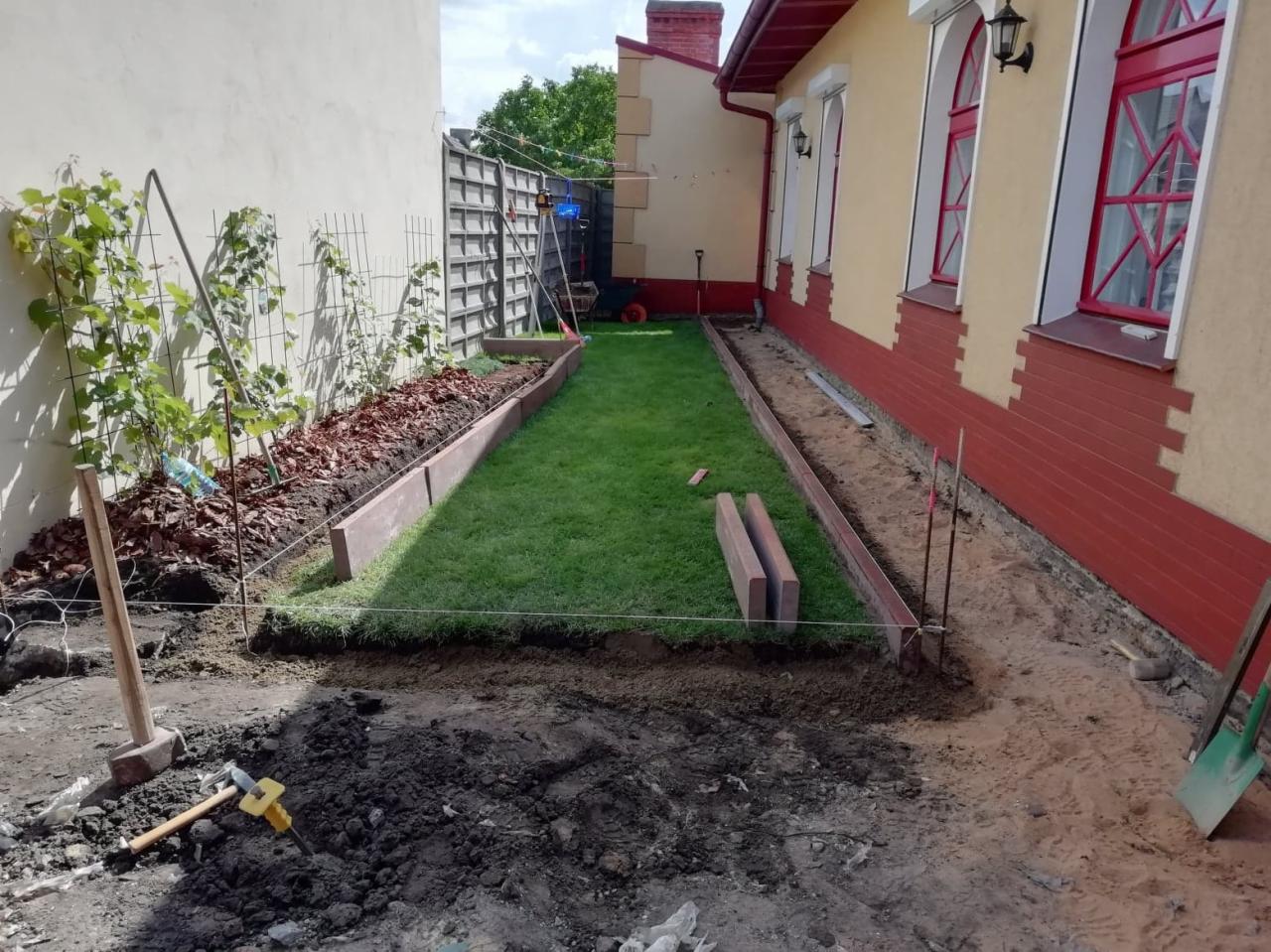 Przygotowanie terenu pod rabaty kwiatowe i trawnik w wąskim pasie ogrodu między budynkami, widoczne obrzeża z kostki, narzędzia ogrodnicze i sznurek wyznaczający granice.