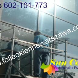 PPUH Sekret-foll - Pracownik na rusztowaniu montuje folię okienną na dużej, przeszklonej fasadzie budynku biurowego; widoczny numer kontaktowy i logo firmy.