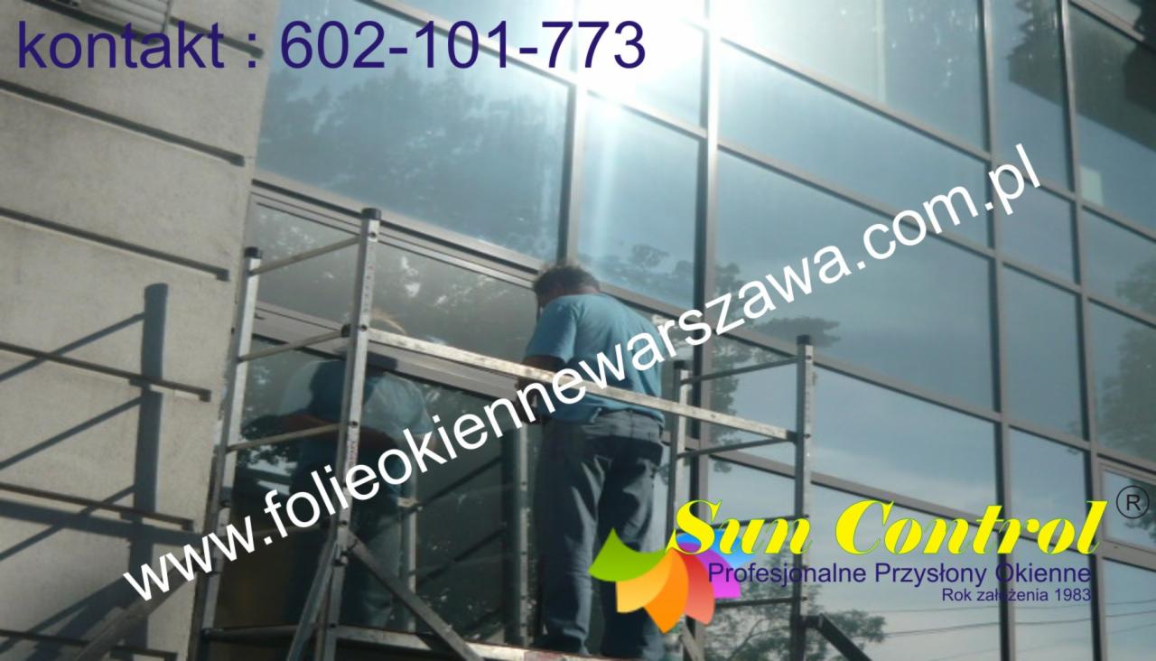 Pracownik na rusztowaniu montuje folię okienną na dużej, przeszklonej fasadzie budynku biurowego; widoczny numer kontaktowy i logo firmy.