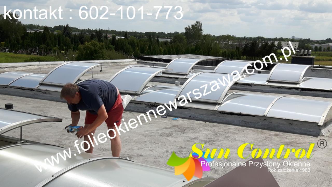 Montaż okien dachowych na płaskim dachu, pracownik uszczelnia silikonem obramowanie, widoczne liczne świetliki dachowe.