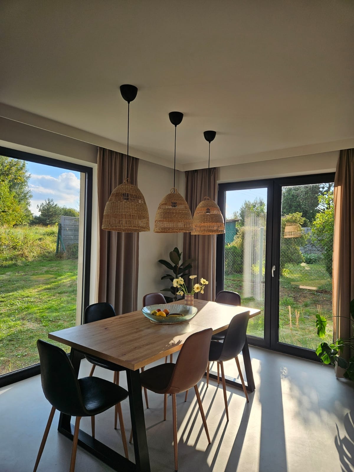 Jadalnia z drewnianym stołem, czterema krzesłami, owocami w misie i trzema wiklinowymi lampami wiszącymi, oświetlona popołudniowym słońcem wpadającym przez duże okna z widokiem na zielony ogród.