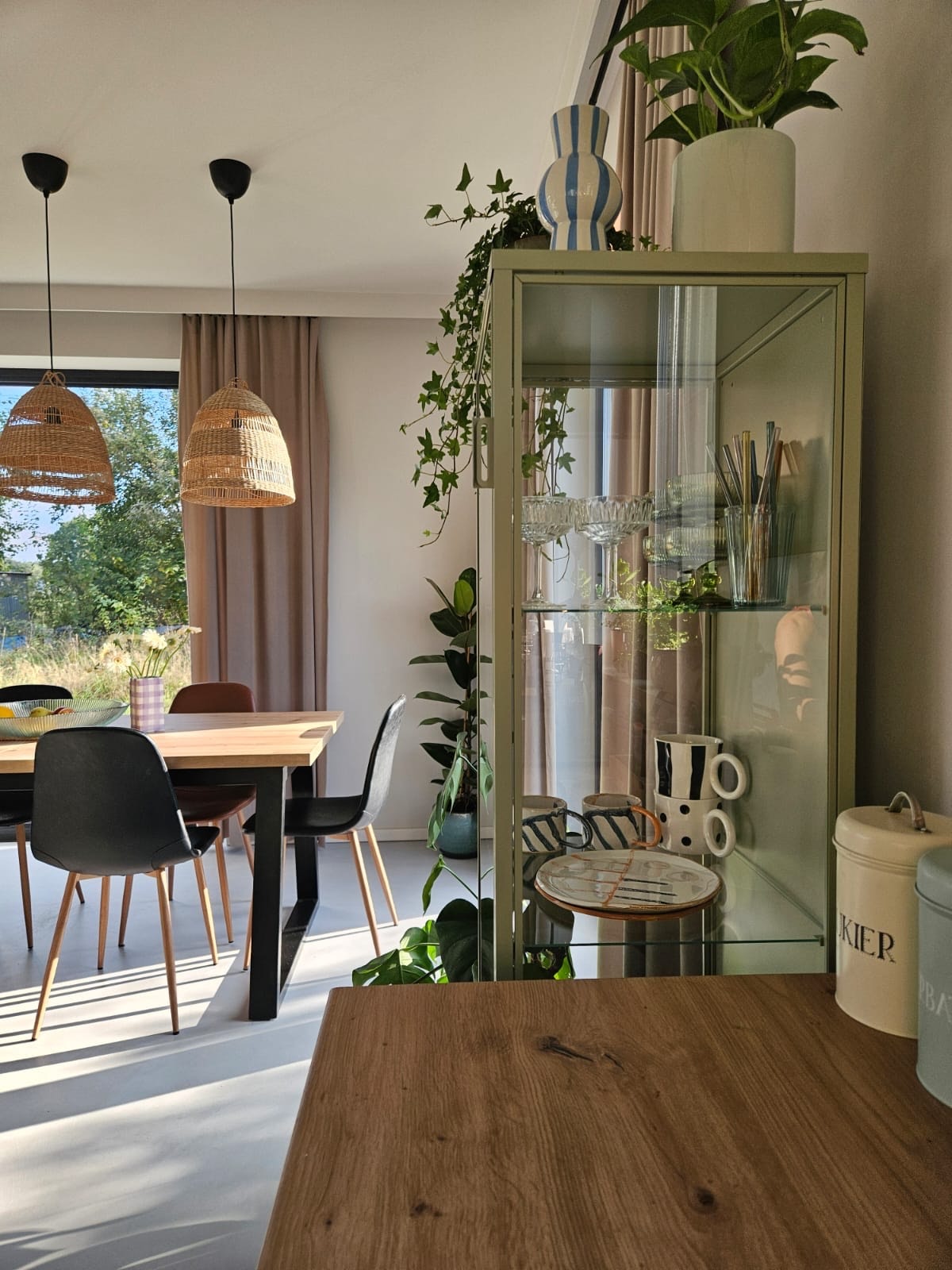 Jasne wnętrze jadalni z drewnianym stołem, czarnymi krzesłami i wiklinowymi lampami. W tle przeszklona witryna w pastelowym kolorze z dekoracyjnymi naczyniami i roślinami.