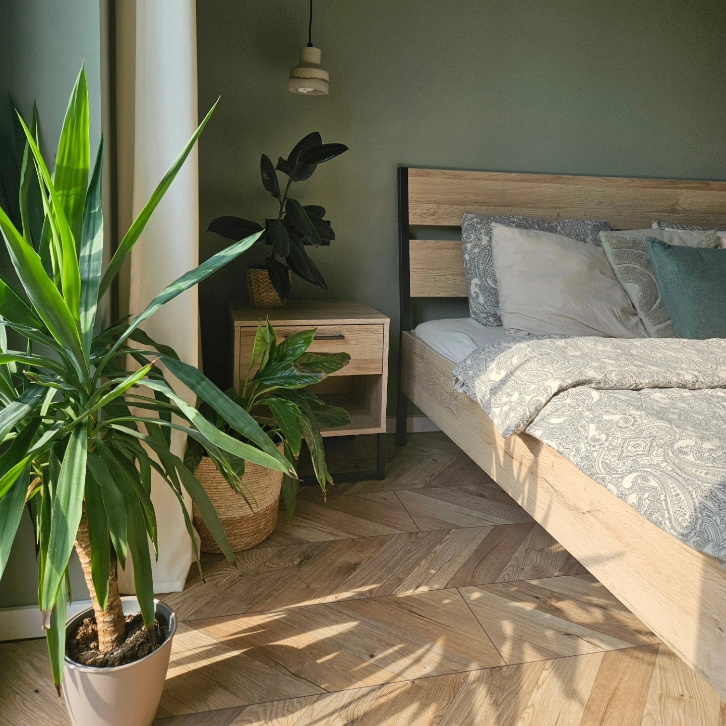 Jasna sypialnia z drewnianą podłogą ułożoną w jodełkę, łóżko z drewnianym zagłówkiem i wzorzystą pościelą, szafka nocna z roślinami doniczkowymi, oliwkowa ściana i wisząca lampa.