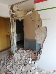 Otwór w ścianie po rozbiórce, widoczne gruzowisko, fragmenty instalacji i warstwy farb na ścianie, surowy stan po demontażu.