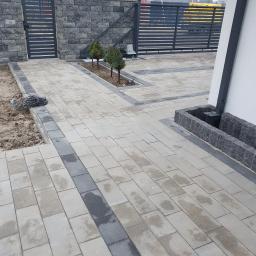 FUGA Brukarstwo Krzysztof Wiśniewski - Ułożona kostka betonowa w odcieniach szarości, obrzeża z ciemniejszej kostki, widoczny fragment ogrodzenia i małe drzewka w wyznaczonym miejscu, kabel elektryczny leżący na ziemi.