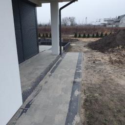 FUGA Brukarstwo Krzysztof Wiśniewski - Nowa nawierzchnia z szarych płyt chodnikowych z ciemnymi obrzeżami, częściowo zasypana piaskiem, prowadząca do domu z szarą roletą i białym murem, w tle widoczna pryzma ziemi i rząd tuj.