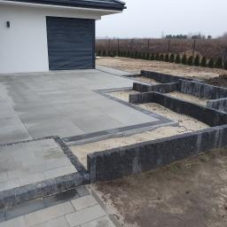 FUGA Brukarstwo Krzysztof Wiśniewski - Ułożona szara nawierzchnia z betonowych płyt przed domem, z obrzeżami z ciemnej kostki. Przygotowane fundamenty pod rabaty kwiatowe z ciemnej kostki, wypełnione piaskiem.