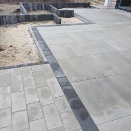 FUGA Brukarstwo Krzysztof Wiśniewski - Fragment posesji z ułożoną szarą nawierzchnią z prostokątnych płyt, obrzeża z ciemnych kostek, przygotowane podłoże pod rabaty z obrzeżami z szarych kostek, widoczne okno budynku w tle.