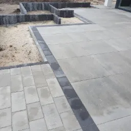 Fragment posesji z ułożoną szarą nawierzchnią z prostokątnych płyt, obrzeża z ciemnych kostek, przygotowane podłoże pod rabaty z obrzeżami z szarych kostek, widoczne okno budynku w tle.