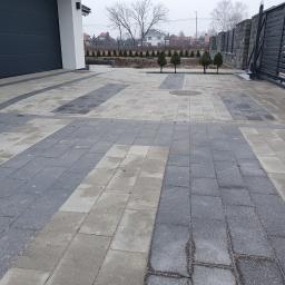 FUGA Brukarstwo Krzysztof Wiśniewski - Dwukolorowa nawierzchnia z kostki brukowej prowadząca od bramy wjazdowej do garażu, z widocznymi młodymi drzewkami w tle i elementami architektury domu.