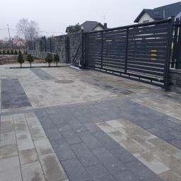 FUGA Brukarstwo Krzysztof Wiśniewski - Ułożona nawierzchnia z kostki w dwóch odcieniach szarości, prowadząca do nowoczesnej, przesuwnej bramy z metalowymi panelami i kamiennego ogrodzenia.