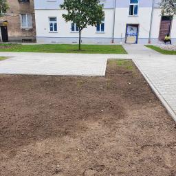 FUGA Brukarstwo Krzysztof Wiśniewski - Nowa, jasnoszara nawierzchnia z kostki na przygotowanym podłożu, z fragmentami trawnika i budynkiem w tle, pracownik w kamizelce siedzi przy wejściu.