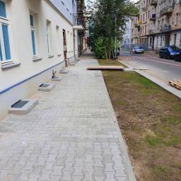 FUGA Brukarstwo Krzysztof Wiśniewski - Nowo ułożony chodnik z szarej kostki brukowej w kształcie litery H, przylegający do budynku z oknami i kratami piwnicznymi, z fragmentem trawnika i ulicy w tle.