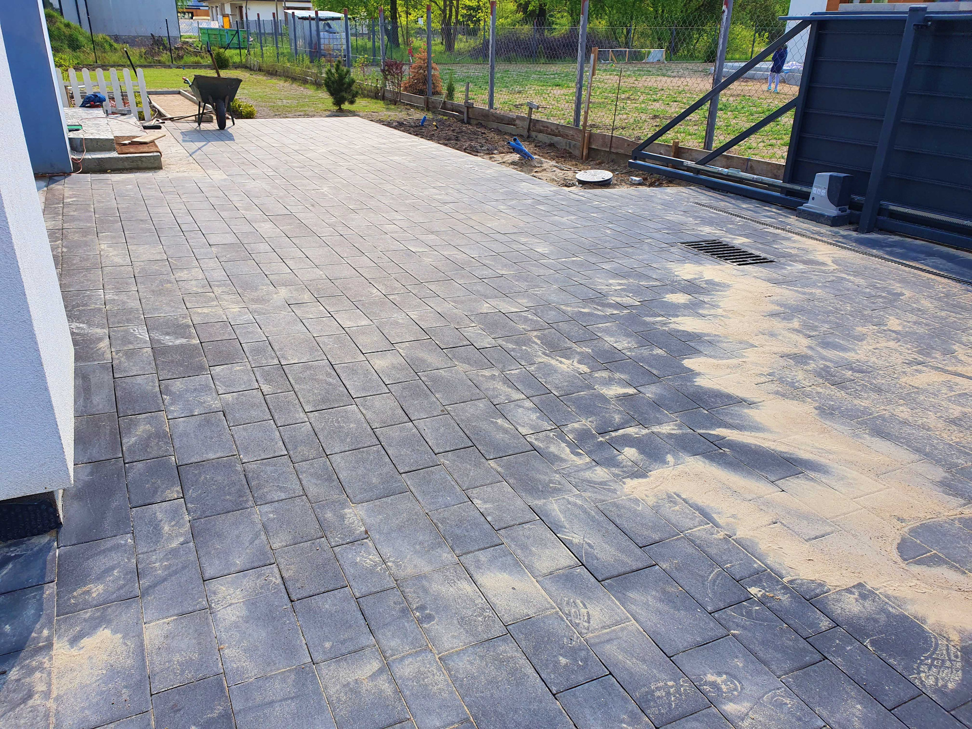 Ułożona, szara nawierzchnia z kostki, częściowo zasypana piaskiem, z widoczną studzienką kanalizacyjną i bramą przesuwną w tle. W oddali widoczny wózek budowlany i białe ogrodzenie.