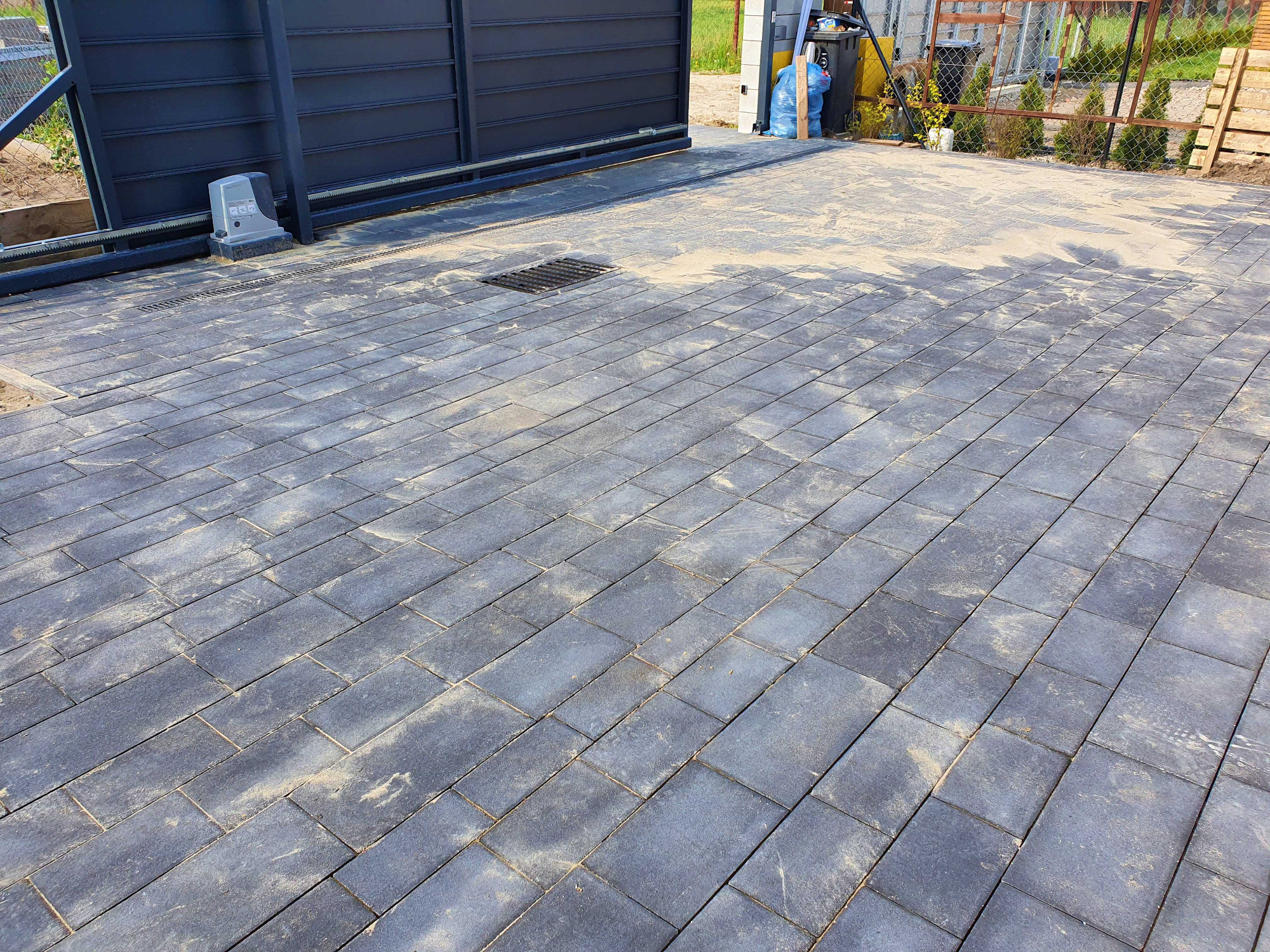 Ułożona szara nawierzchnia z prostokątnych elementów przy automatycznej bramie wjazdowej, widoczny odpływ liniowy.