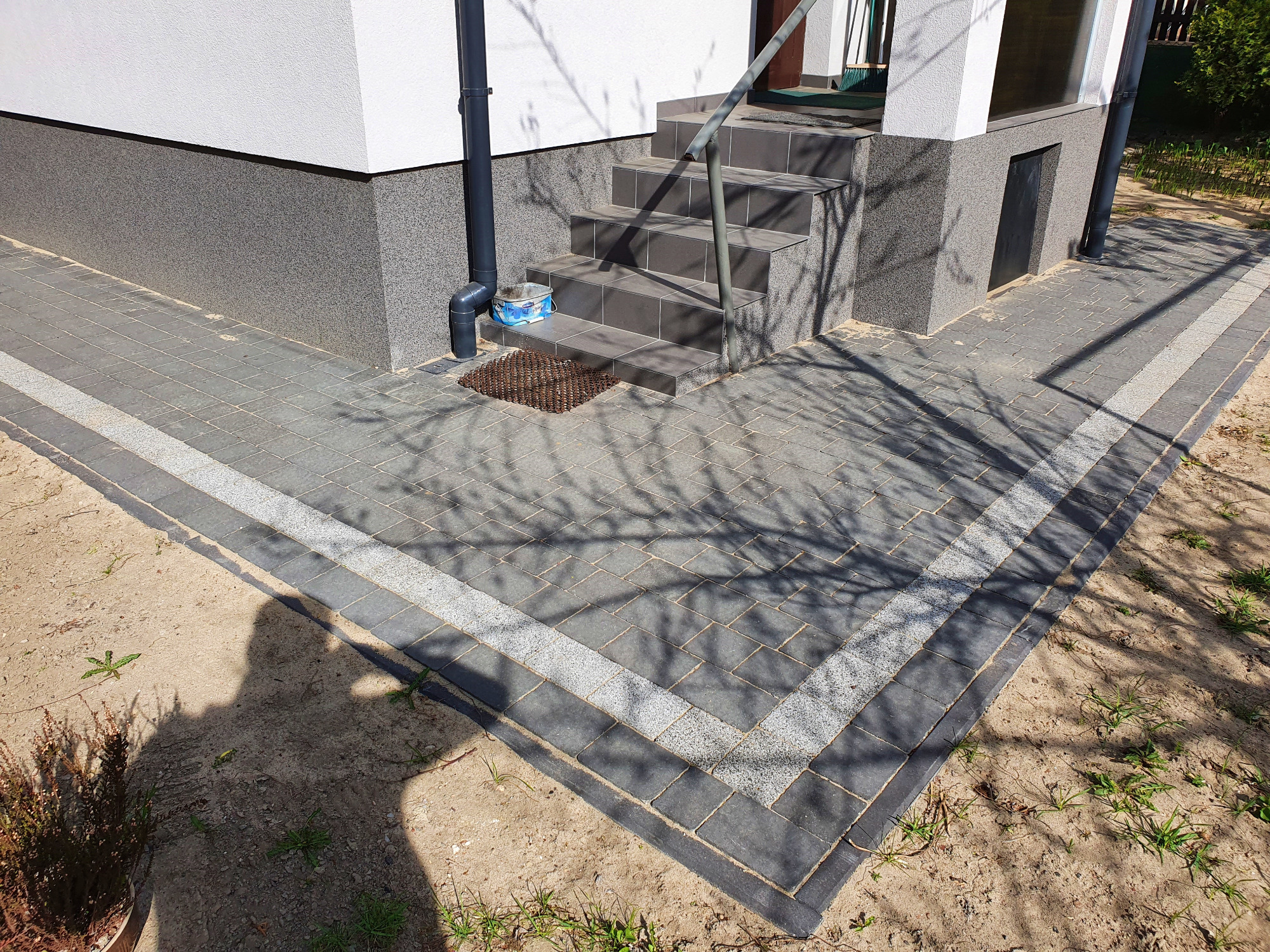Ułożona szara kostka brukowa z jasnoszarym obramowaniem przed wejściem do domu z szarymi schodami i metalową poręczą, widoczny cień osoby.