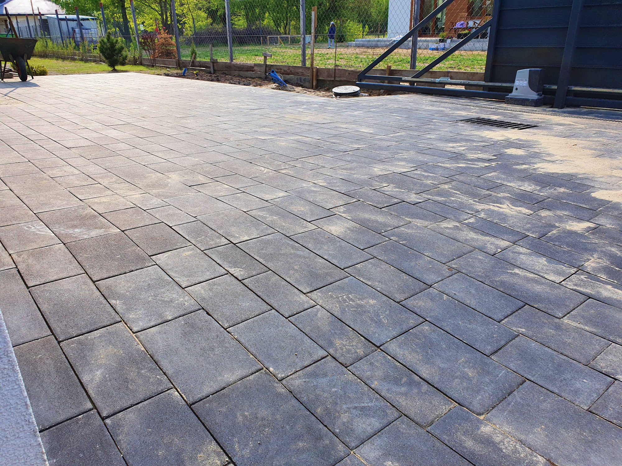 Ułożona, prostokątna kostka brukowa w odcieniach szarości na podjeździe, z widoczną bramą w tle i fragmentem ogrodu.