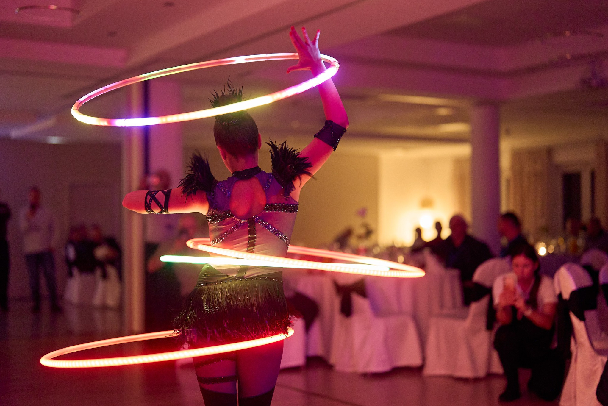 Kobieta w kostiumie z piórami prezentuje pokaz z podświetlanymi hula-hoop na tle sali bankietowej z gośćmi. Scena oświetlona różowym światłem.