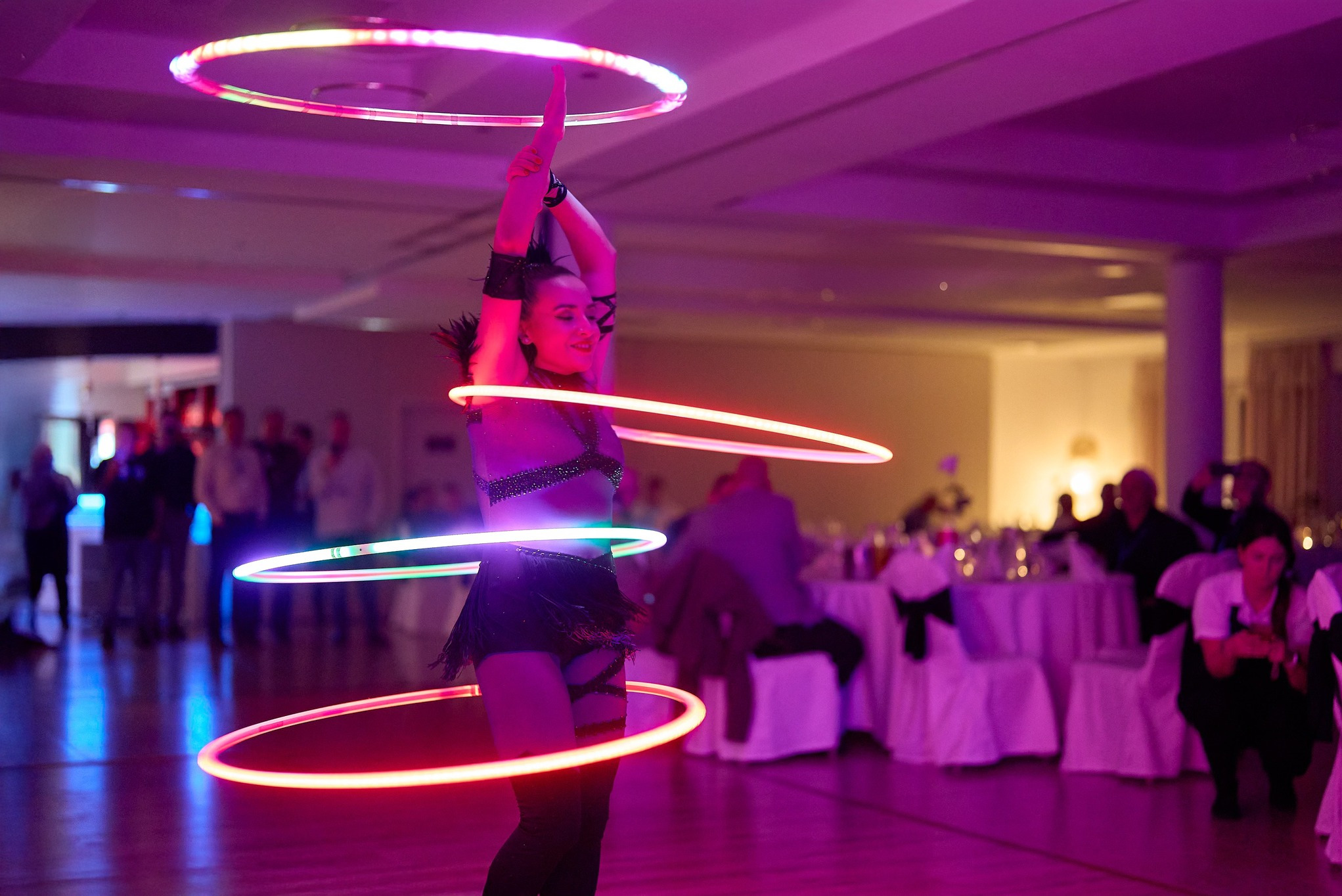 Artystka z hula-hoop LED na scenie podczas eventu. Dynamiczne światła i ruch tworzą efektowny pokaz w fioletowej tonacji. Widoczni goście w tle.