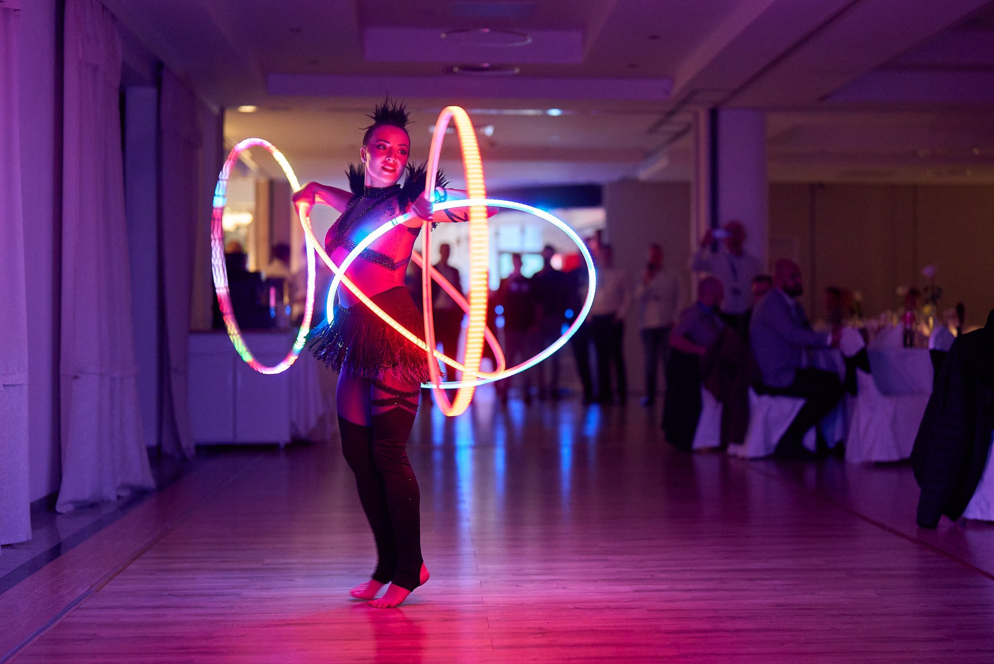 Kobieta w kostiumie prezentuje pokaz z podświetlanymi hula-hoop na parkiecie. W tle widoczni goście siedzący przy stołach. Scena oświetlona fioletowym światłem.