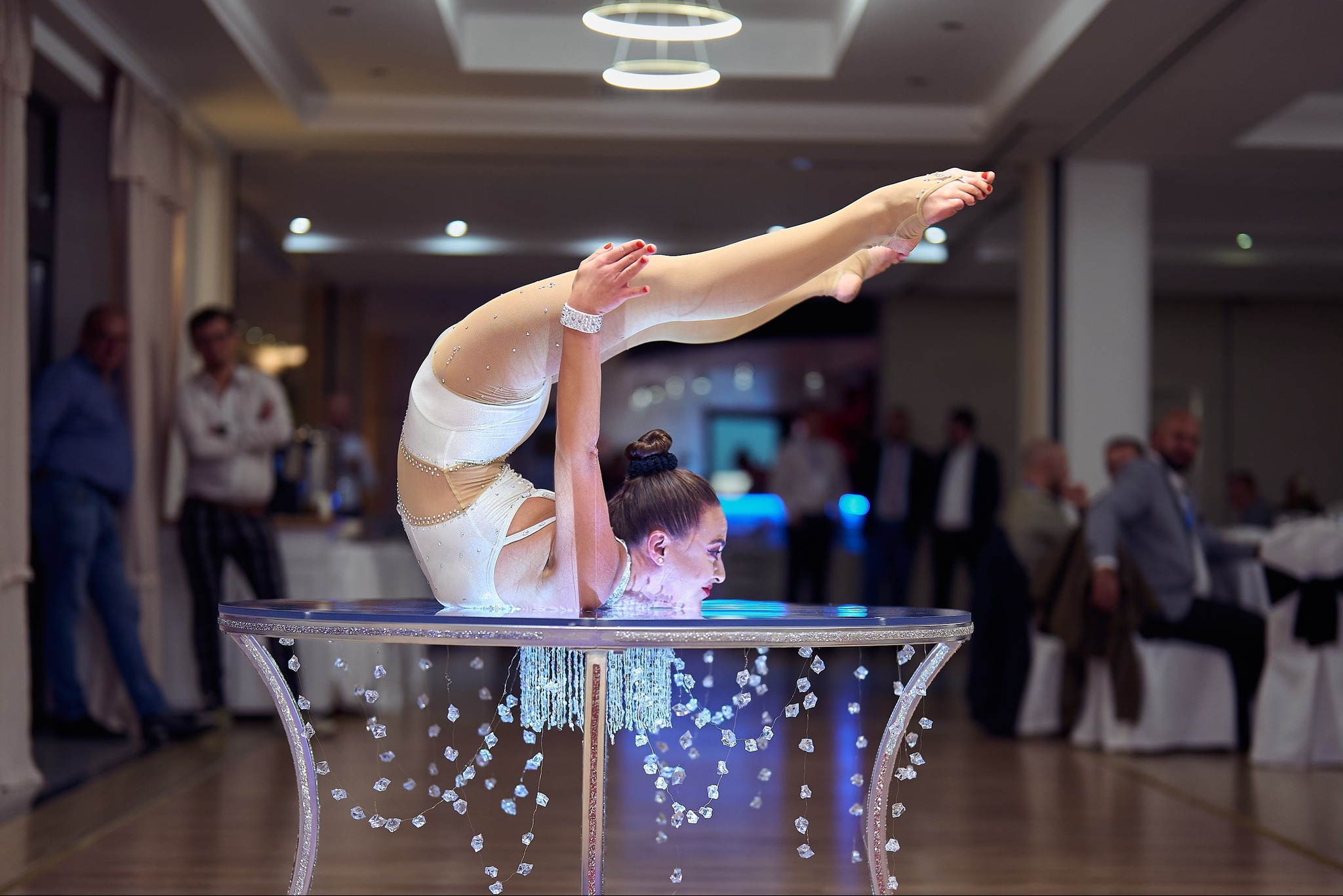 Akrobatka w białym kostiumie prezentuje ekstremalne wygięcie na dekoracyjnym stole z kryształkami podczas występu na tle sali bankietowej.