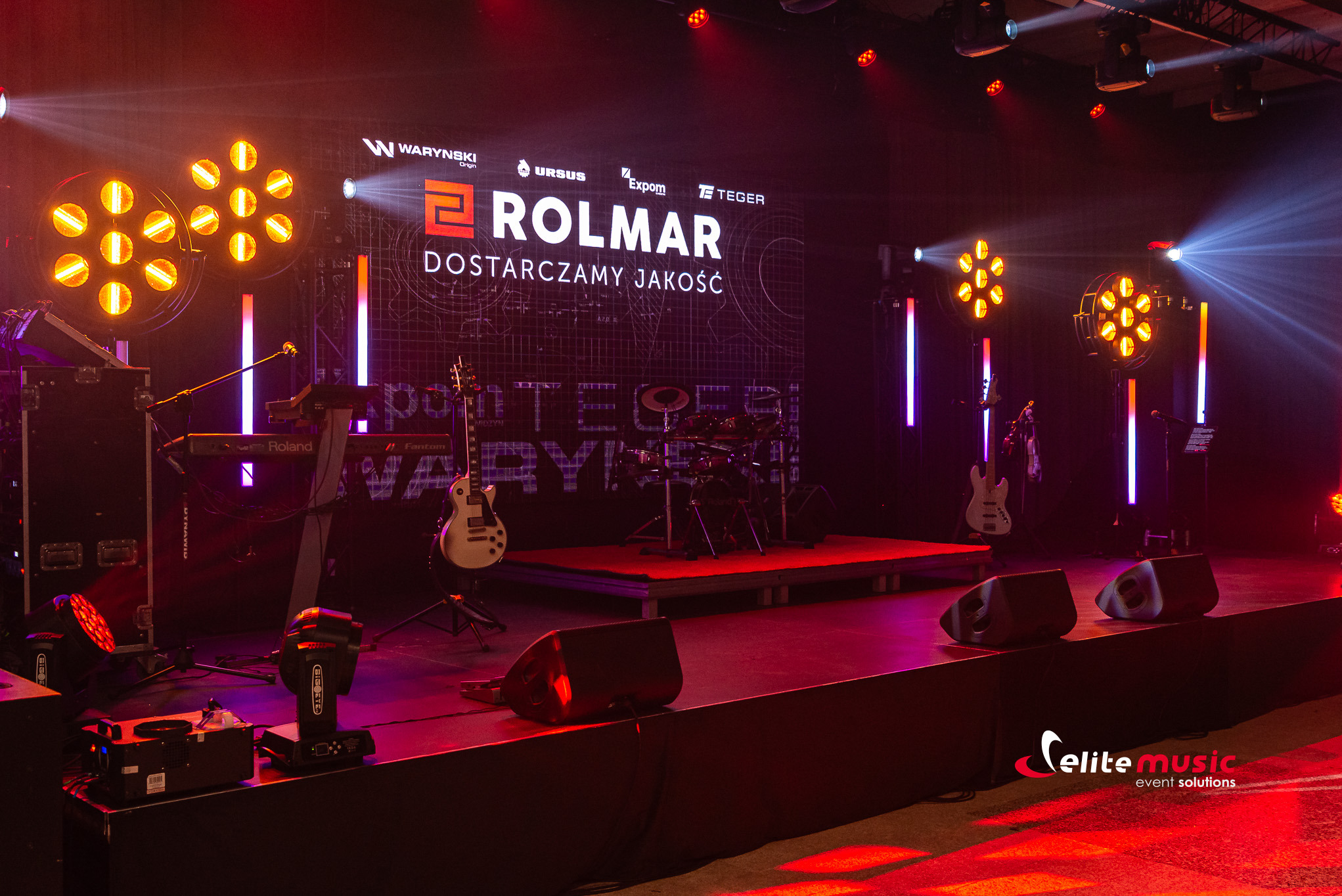 Scena eventowa z instrumentami (gitara, perkusja, keyboard) i oświetleniem. Na tylnej ścianie logo firmy Rolmar. Widoczne głośniki i efekty świetlne.