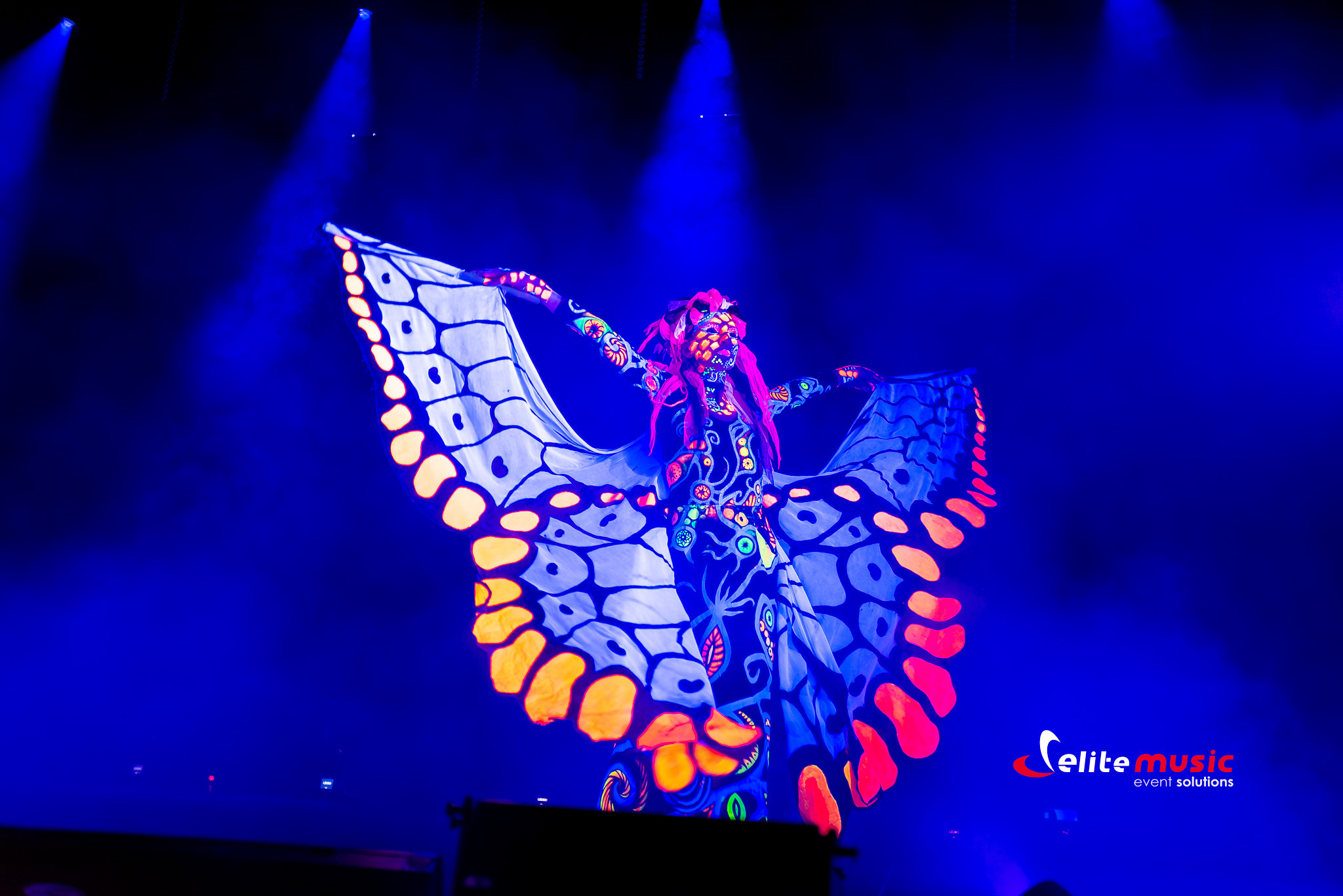 Artystka w kostiumie motyla UV na scenie, oświetlona niebieskim światłem. Fluorescencyjne wzory na ciele i skrzydłach. Pokaz UV Show podczas eventu.