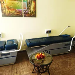 Dwa specjalistyczne fotele rehabilitacyjne z panelami sterowania, obok stolik z owocami, w tle obraz i regał z hantlami w gabinecie.