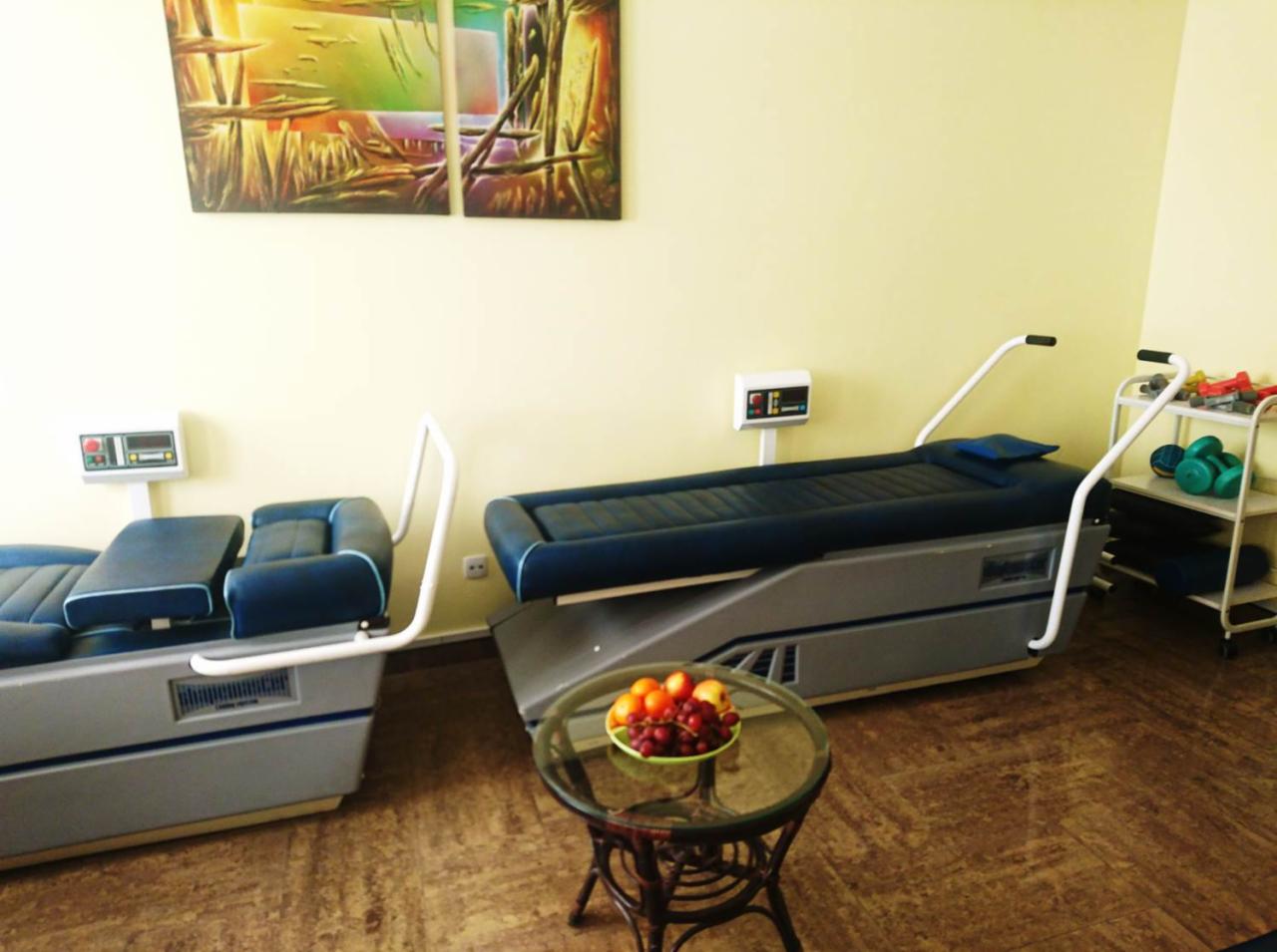 Dwa specjalistyczne fotele rehabilitacyjne z panelami sterowania, obok stolik z owocami, w tle obraz i regał z hantlami w gabinecie.