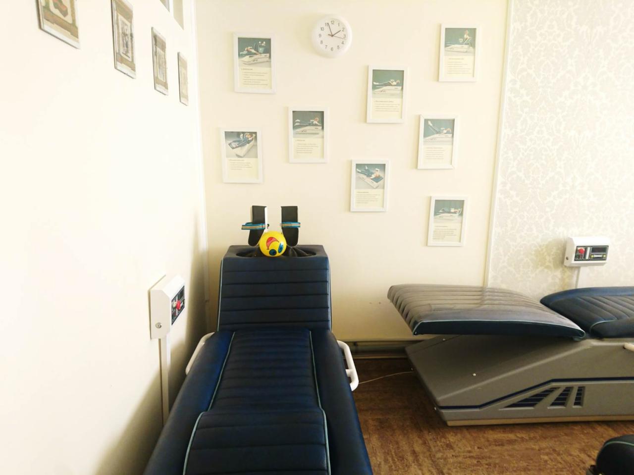 Gabinet rehabilitacyjny z dwoma specjalistycznymi łóżkami do terapii kręgosłupa, ozdobiony ramkami ze zdjęciami i zegarem na ścianie.