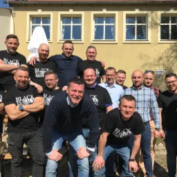 Grupa mężczyzn w koszulkach z napisem 'Black is Back Specialist 1872' pozuje na zewnątrz budynku z żółtą elewacją i białymi oknami, prawdopodobnie po zakończonym szkoleniu lub evencie firmowym.