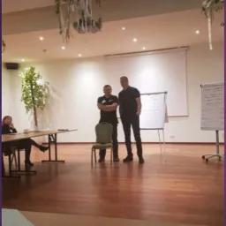 Prelegenci podczas szkolenia w Grodzisku Wielkopolskim, stojący przed tablicą flipchart. W tle widoczna siedząca osoba przy stole i dekoracje sali.