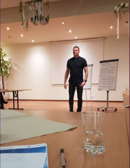 Mężczyzna w czarnej koszulce i spodniach stojący w sali konferencyjnej przed flipchartem z notatkami, widok zza stołu z długopisem i szklanką wody.