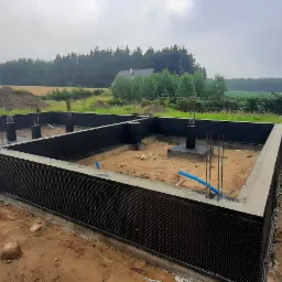 Fundament domu murowanego z izolacją pionową i wystającymi prętami zbrojeniowymi, pracownik wyr&oacute;wnuje teren wok&oacute;ł fundamentu, krajobraz wiejski w tle.