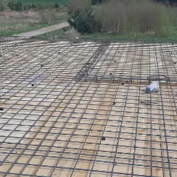 Zbrojenie płyty fundamentowej z siatki stalowej na drewnianym szalunku, widoczne elementy izolacji i worki z materiałami budowlanymi w tle